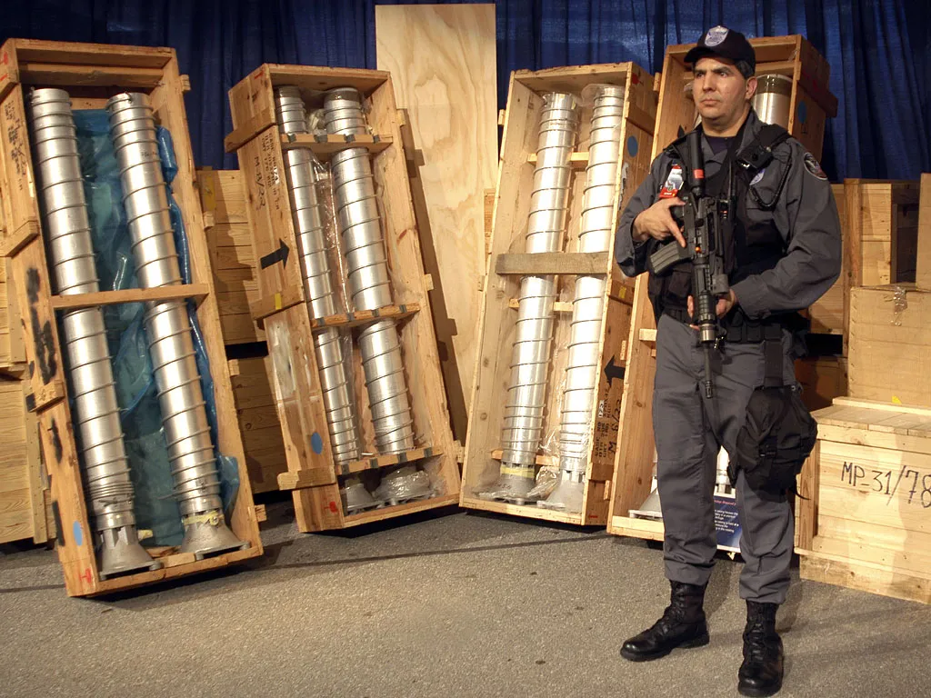 A security officer stands armed next to large metal pipes packed in wooden crates, displayed upright against a blue curtain backdrop.