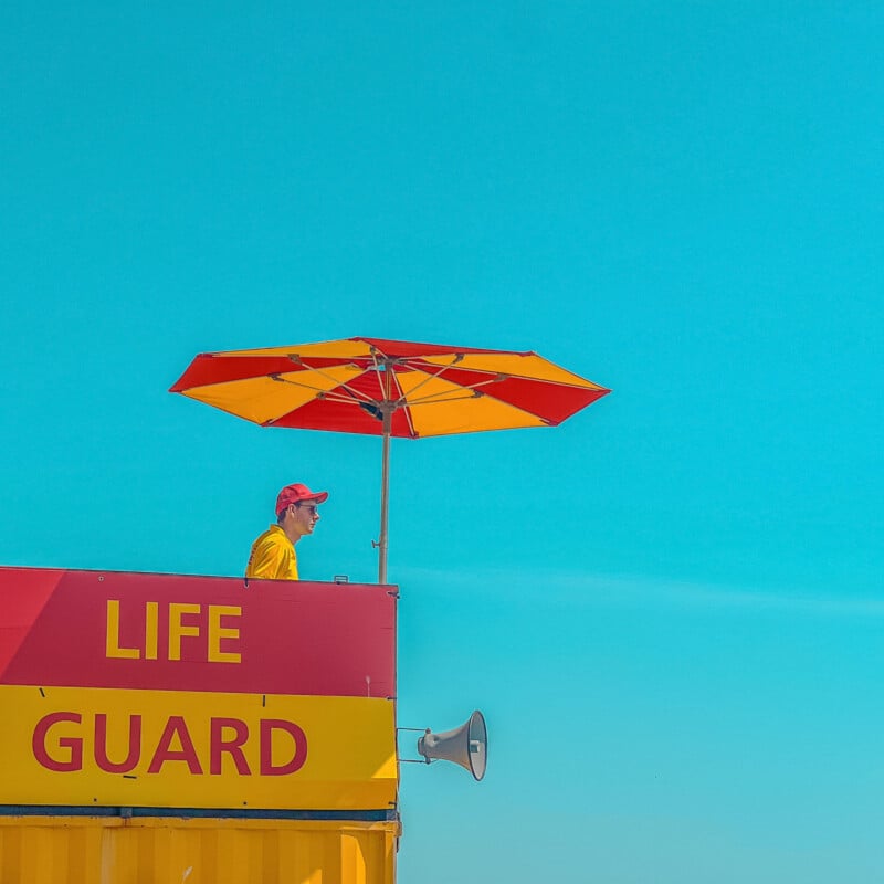 A lifeguard wearing a red cap and yellow shirt sits under a red and yellow umbrella atop a lifeguard station, with a bright blue sky in the background.