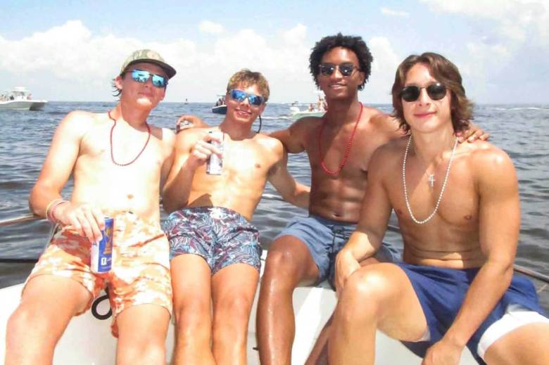 Four young men in swim trunks, three white and one black, pose together on the back of a small boat on the water. They are holding beer cans.