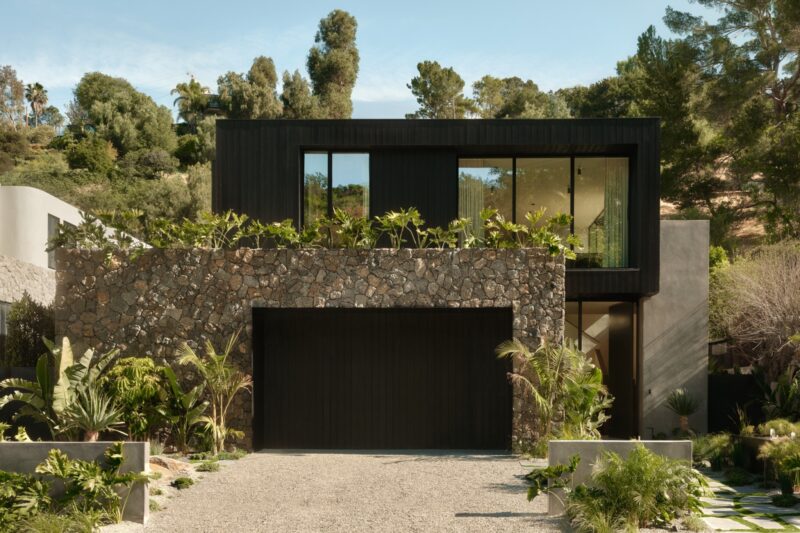 Modern two-story Obsidian House with large windows, stone facade on the lower level, black upper level, surrounded by lush green plants and trees. Gravel driveway leads to a black garage door.