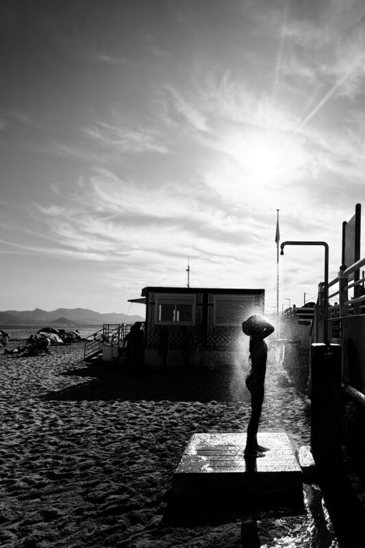 A person showering on a beach in silhouette, with sunlight streaming through misted water and buildings, sand, and distant mountains visible in the background.