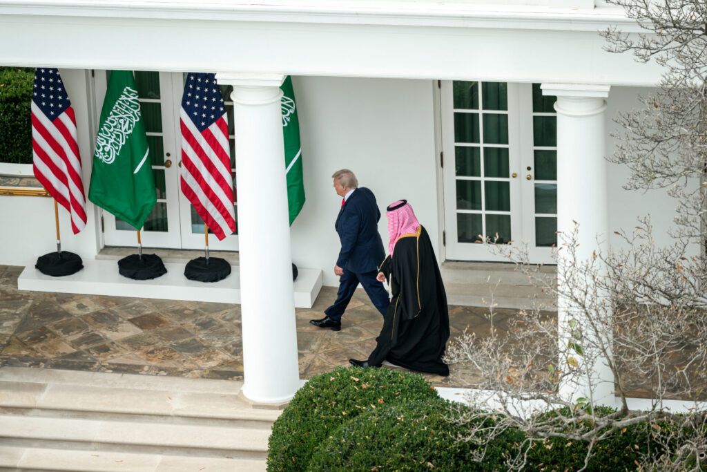 Two men walk outside a white building past American and Saudi Arabian flags; one wears a suit and the other wears traditional Middle Eastern attire.