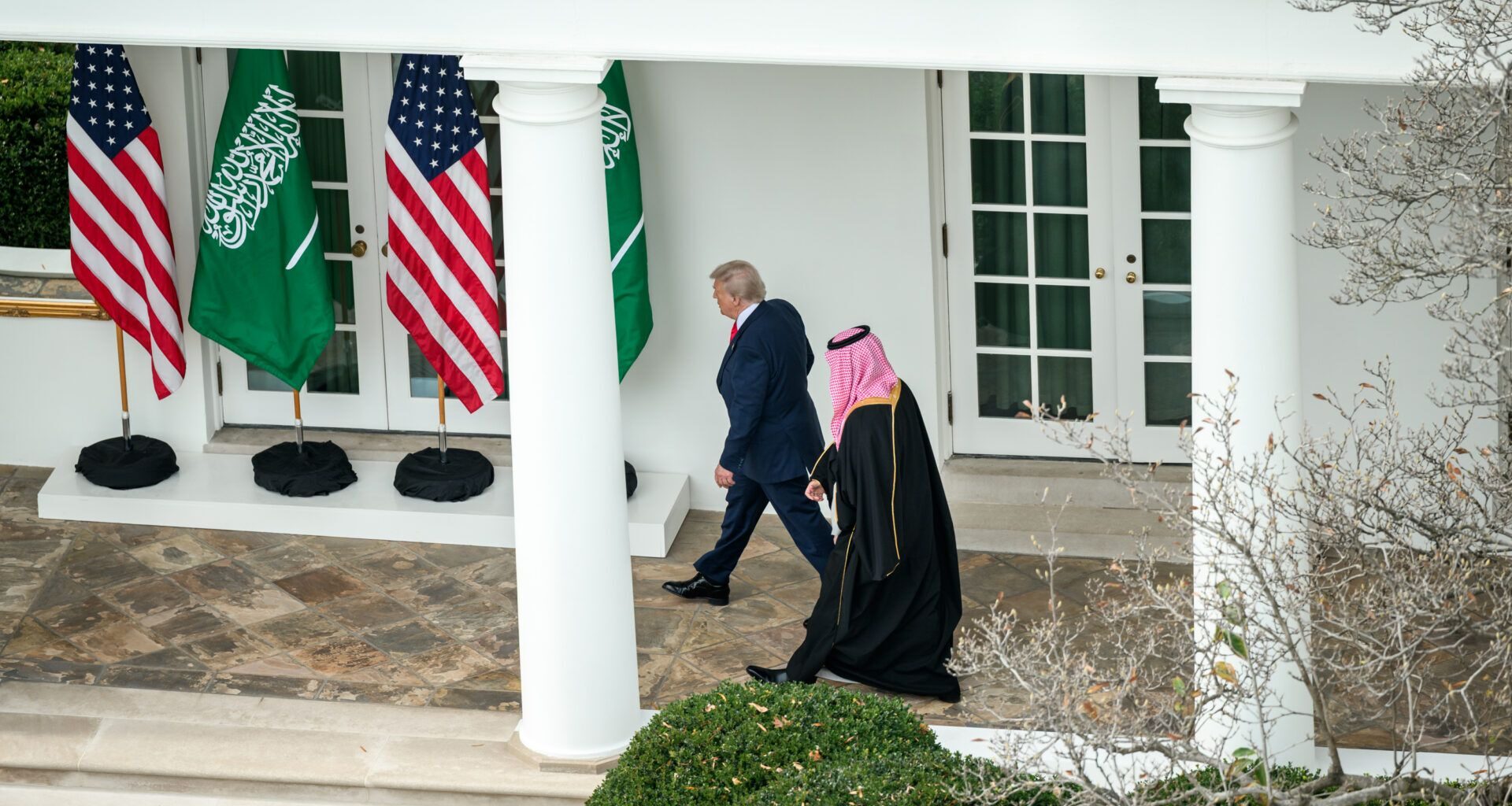 Two men walk outside a white building past American and Saudi Arabian flags; one wears a suit and the other wears traditional Middle Eastern attire.