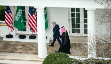 Two men walk outside a white building past American and Saudi Arabian flags; one wears a suit and the other wears traditional Middle Eastern attire.