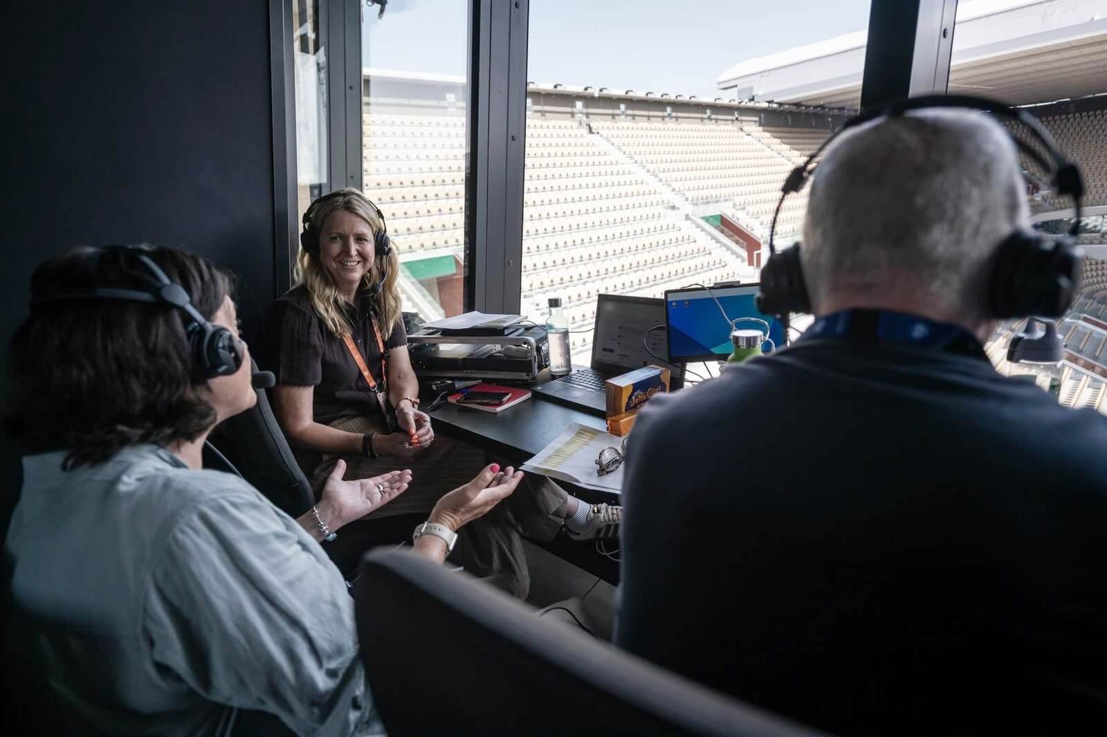Gigi Salmon (left) sits in a commentary box wearing over-ear headphones with two other commentators doing the same, who have their backs to the camera. The window looks out on to an empty Court Philippe-Chatrier stand, at the French Open.