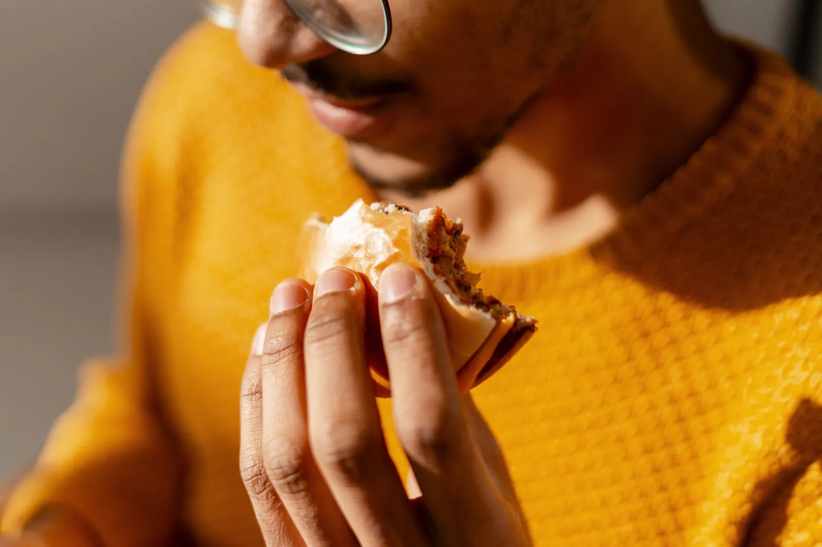 A man holding and eating a hamburger, wearing glasses and yellow sweater.