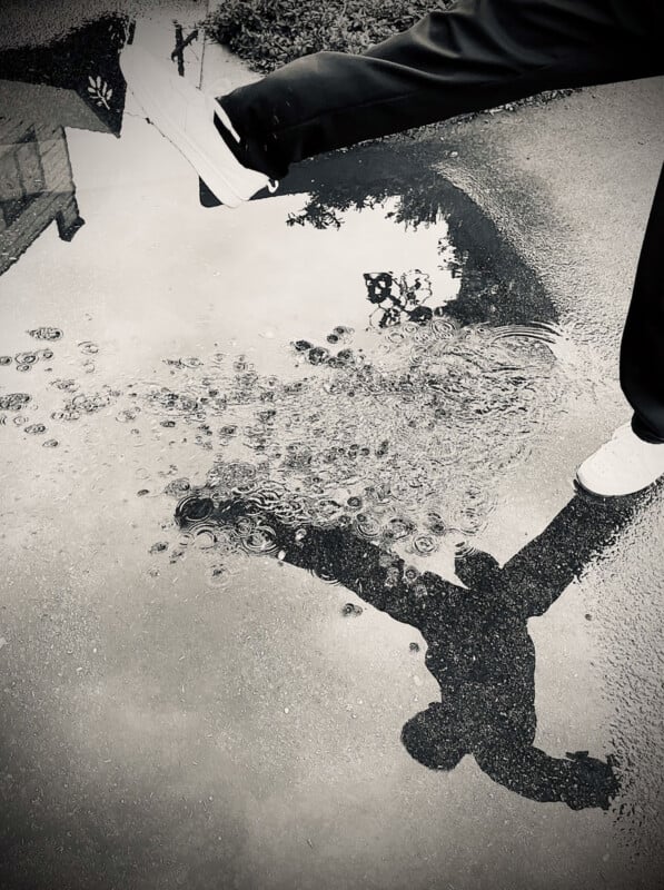 A black and white photo of a person stepping into a puddle, creating ripples. The person's shadow is clearly reflected in the water, while only their legs and white shoes are visible above the ground.