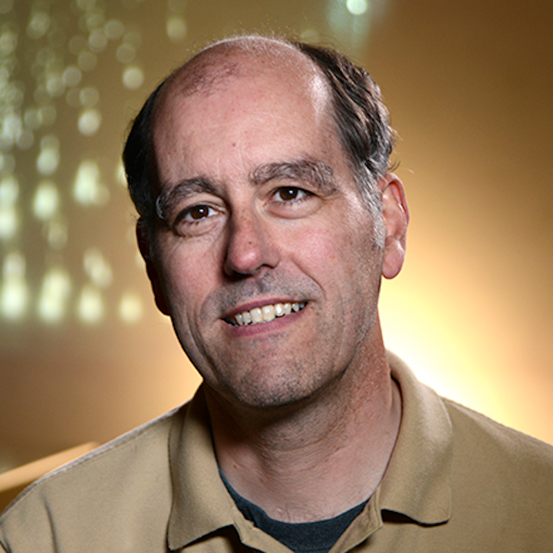 Partly bald smiling man wearing a tan shirt.