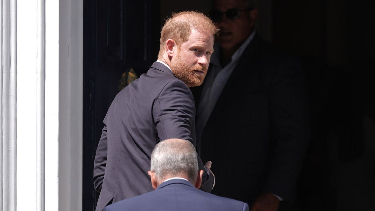Prince Harry in a dark suit looking from his shoulder as a bodyguard walks in front of him,