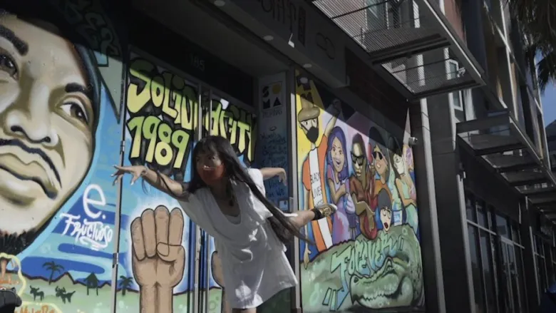 A person poses on one leg in front of a colorful mural featuring portraits, a raised fist, and text reading "SOLU WIDGETS 1989.