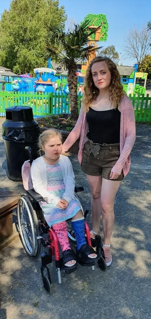 A woman stands next to a young girl in a wheelchair, who wears casts on both legs.