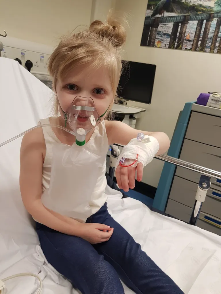 A young girl wearing an oxygen mask and with an IV in her arm, sitting on a hospital bed.