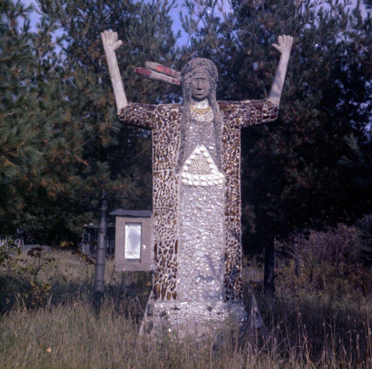 A stone and mosaic statue of a Native American figure with raised arms stands in a grassy area, surrounded by trees.