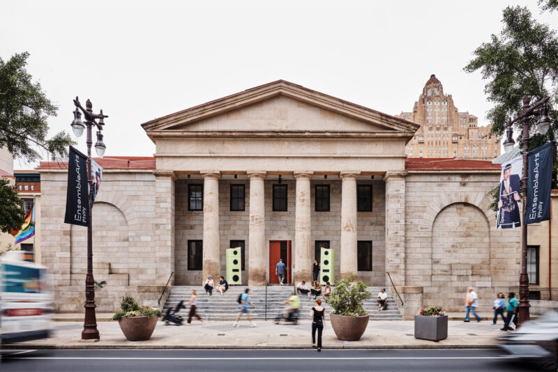 Neoclassical building with tall columns and stone facade, people walking on the sidewalk and steps, banners and trees on both sides, city buildings in background.