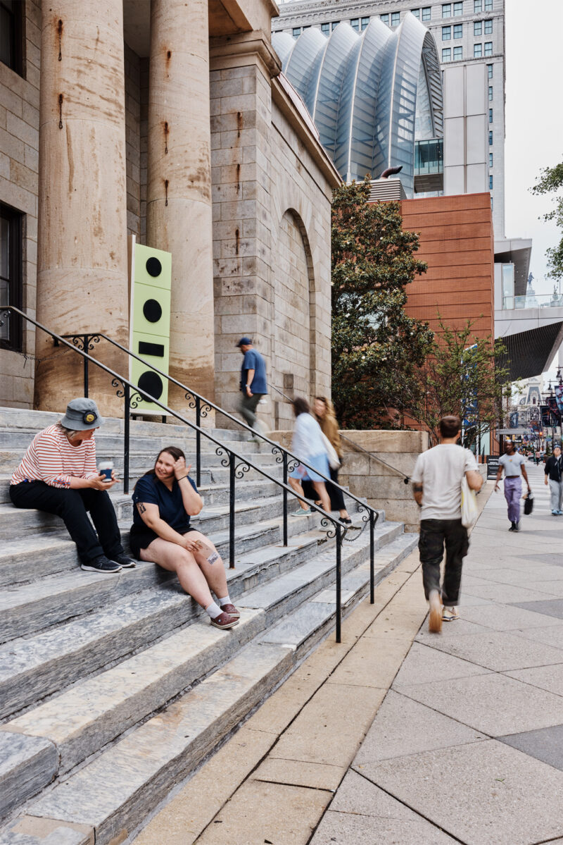 People sit and talk on stone steps outside a large building, while others walk along the sidewalk; modern buildings are visible in the background.