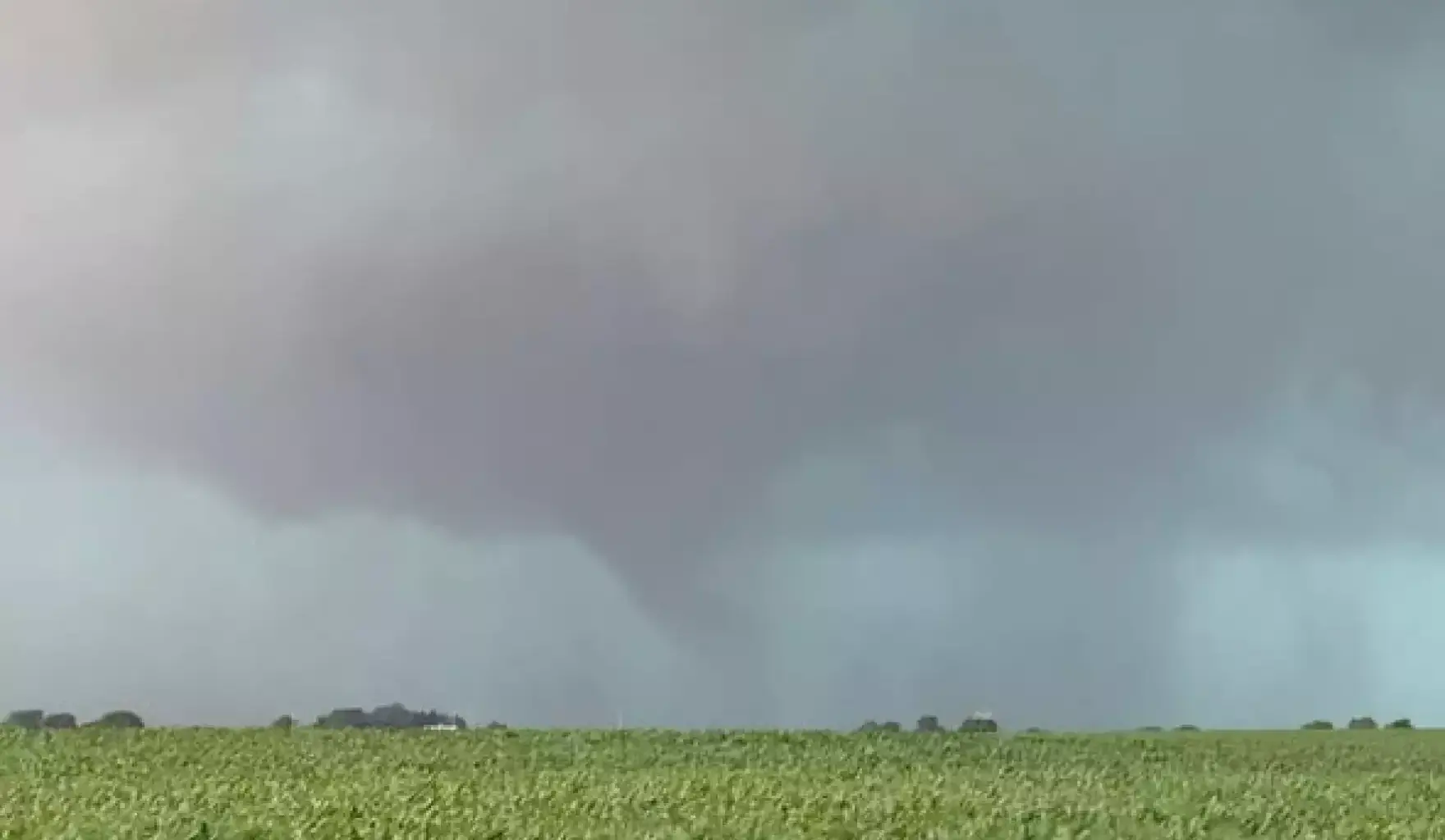 A funnel cloud extends from a dark, stormy sky above a green field, indicating the early formation of a tornado.