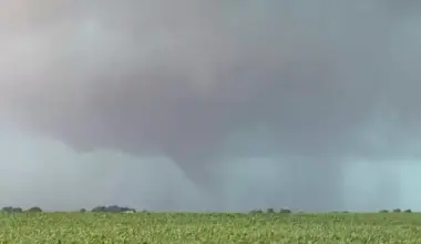 A funnel cloud extends from a dark, stormy sky above a green field, indicating the early formation of a tornado.