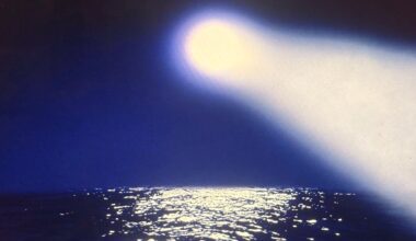 A bright light shines down from the sky onto a dark ocean, creating a glowing reflection on the water's surface under a deep blue night sky.