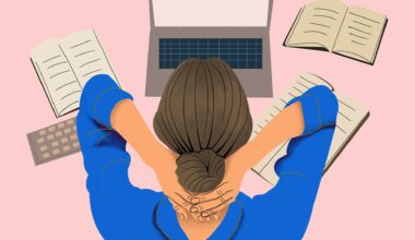 High angle view of a female office worker with hands around back of neck with laptop and books scattered around a desk.