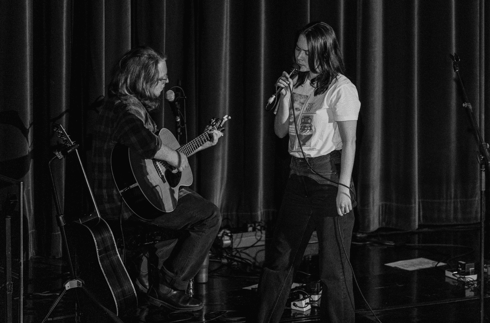 A musician plays guitar while a singer performs on stage under soft lighting.
