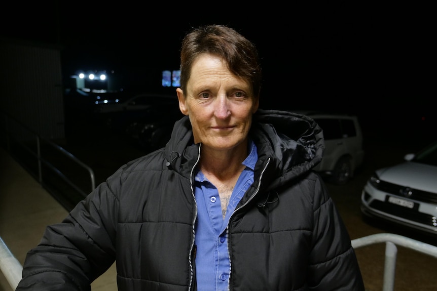 Sue with short hair gazes directly at the camera, background is dark, cars parked.