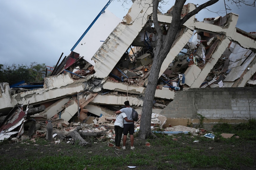 A man and woman embracing, seen from behind, next to a tree and in front of a collapsed building.