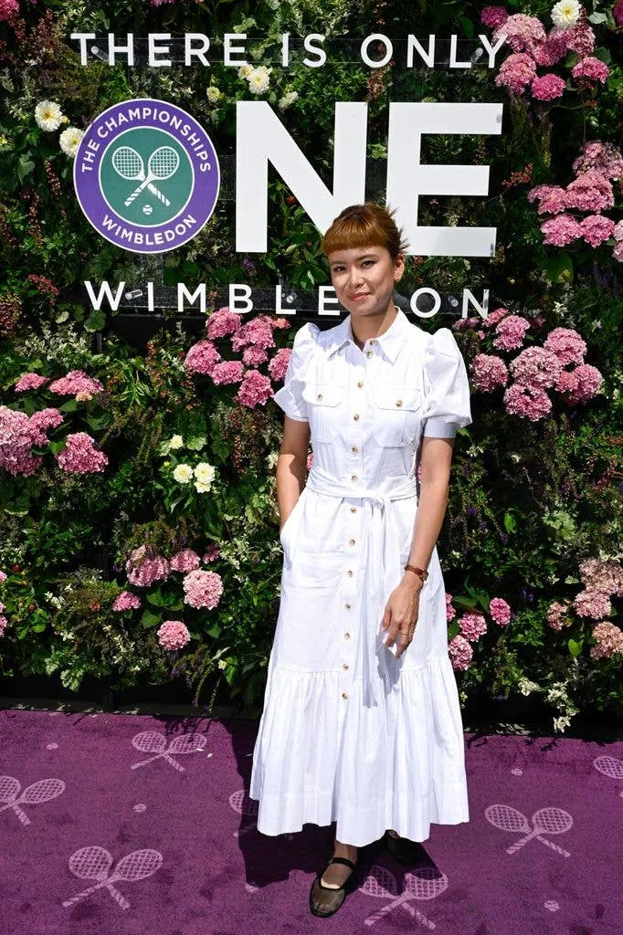 Katie Leung stands in front of a floral backdrop, wearing a white dress and smiling.