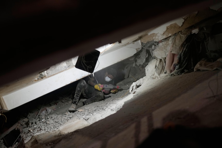 A man in dark clothing and a face mask, lying under a slab of concrete in a sliver of light next to a machine jack.