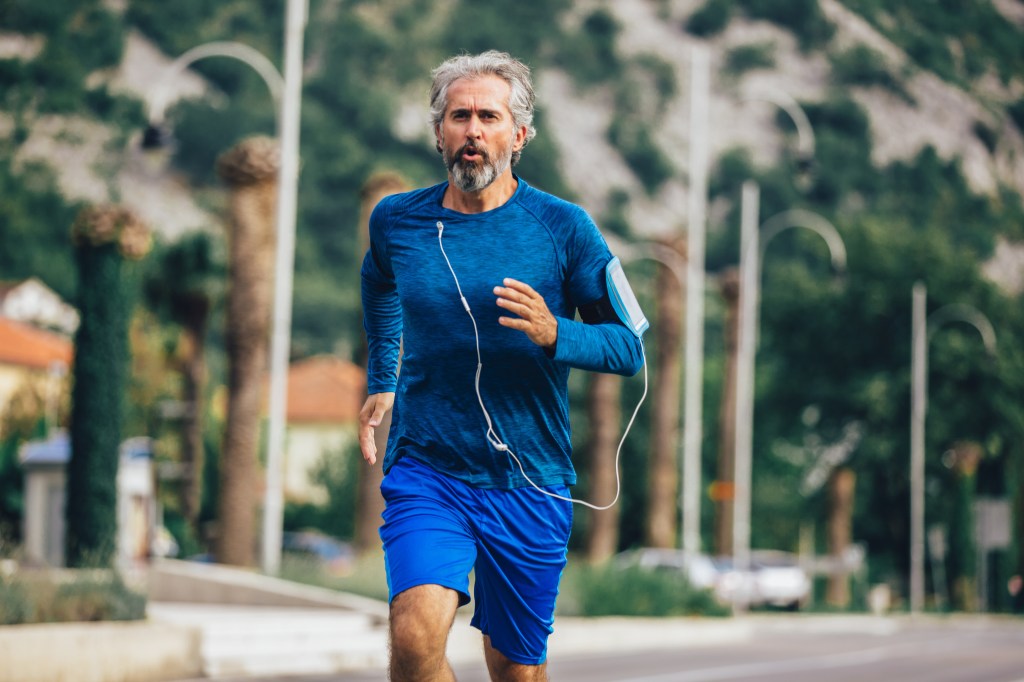 An active senior man with gray hair and a beard jogs on a sunny day, wearing a blue shirt and shorts, with earphones and a phone armband.