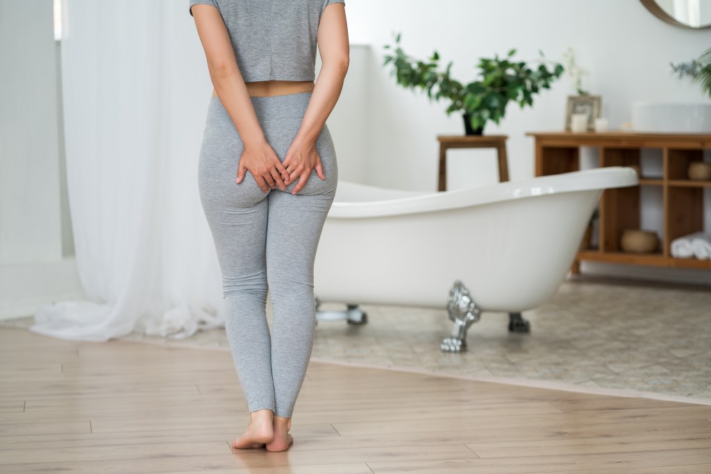 A woman suffering from hemorrhoids holds her hands to her bottom in a bathroom.