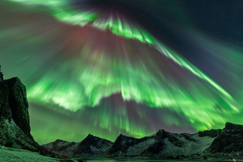Bright green and purple aurora borealis lights stretch across a starry night sky above dark, jagged mountains and a snowy landscape.