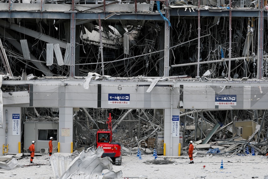Rescue workers in orange stand next to a digger machine amid the rubble of a building.