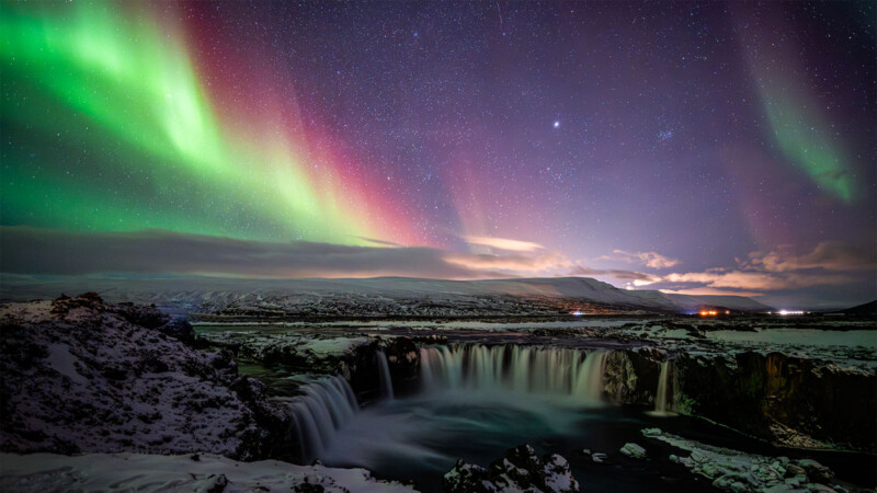 A snowy landscape with a waterfall under a starry night sky, illuminated by vibrant green and pink aurora borealis lights.