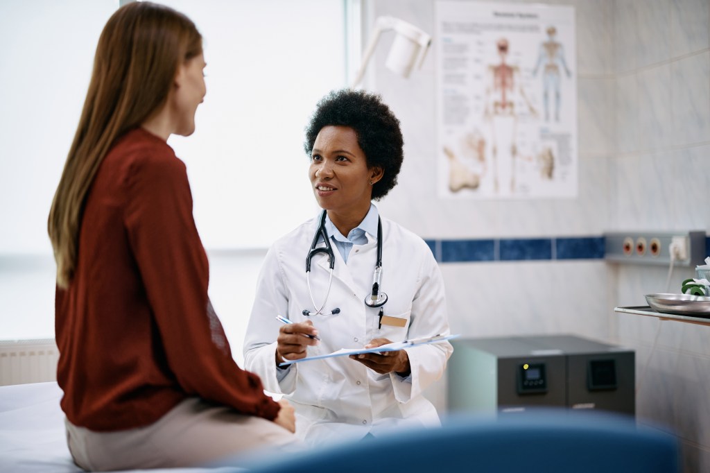 An African American female doctor in a white coat talks to a female patient during a medical exam.