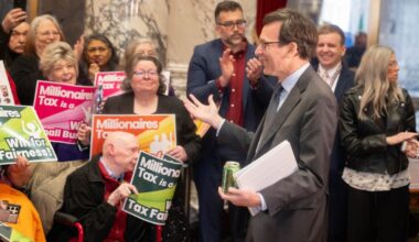 Gov. Bob Ferguson gestures to a crowd of supporters holding "Millionaires Tax is a Win for Tax Fair...