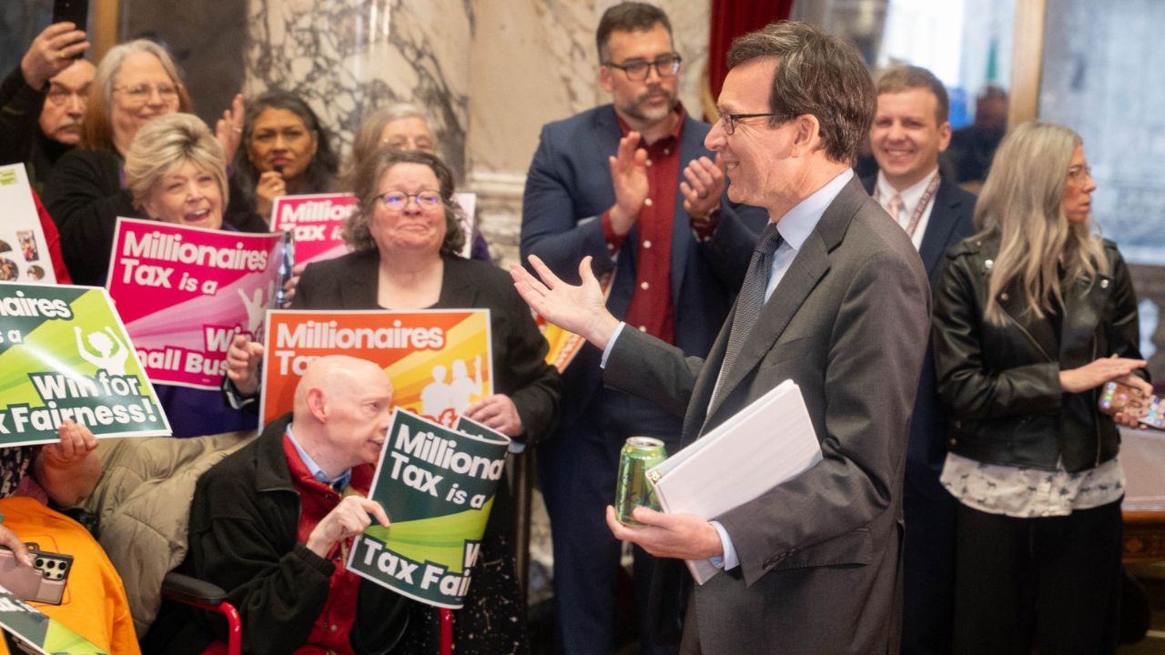 Gov. Bob Ferguson gestures to a crowd of supporters holding "Millionaires Tax is a Win for Tax Fair...
