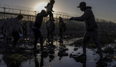 Migrants silhouetted against the sunrise as they cross a muddy riverbed near a razor-wire border fence.