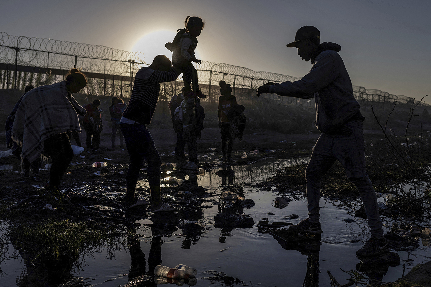 Migrants silhouetted against the sunrise as they cross a muddy riverbed near a razor-wire border fence.