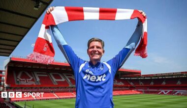 Oliver Glasner faces into the sunshine holding a red and white bar scarf, with the City Ground pitch and stands in the background