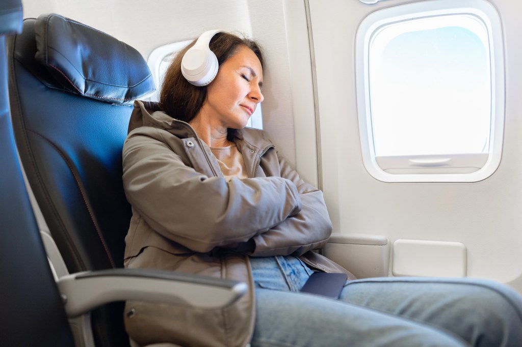 Woman in a gray jacket and white headphones sleeping peacefully by an airplane window.