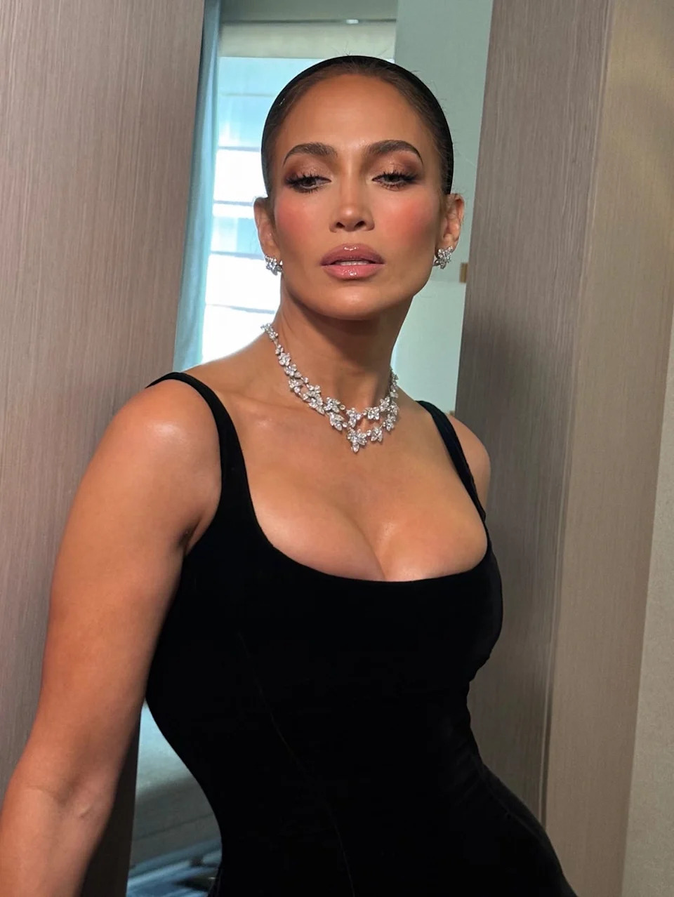 A woman in a black dress and sparkling jewelry poses confidently in a well-lit room.