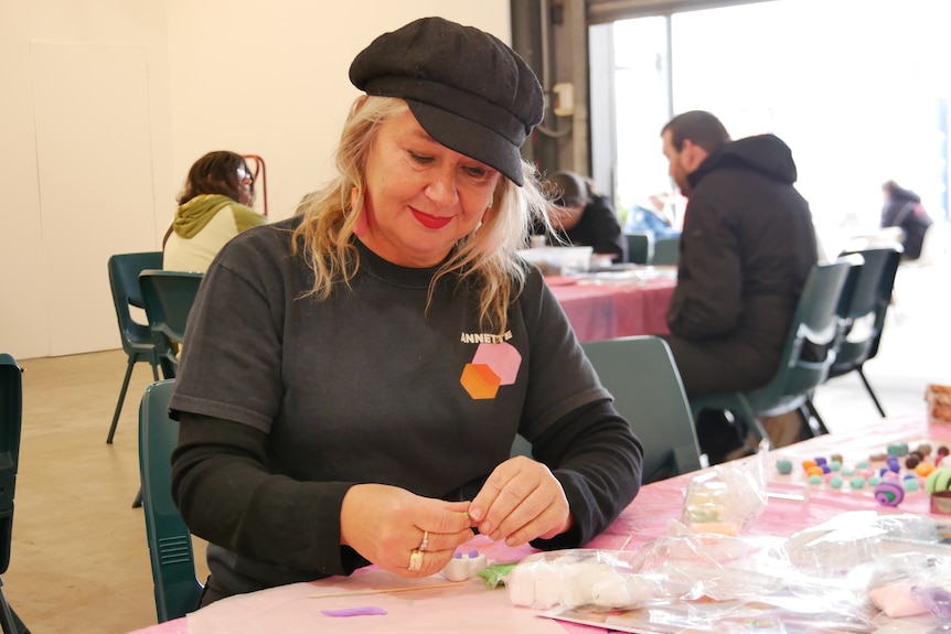 A woman wearing a black hat sits at a table hand making clay beads.