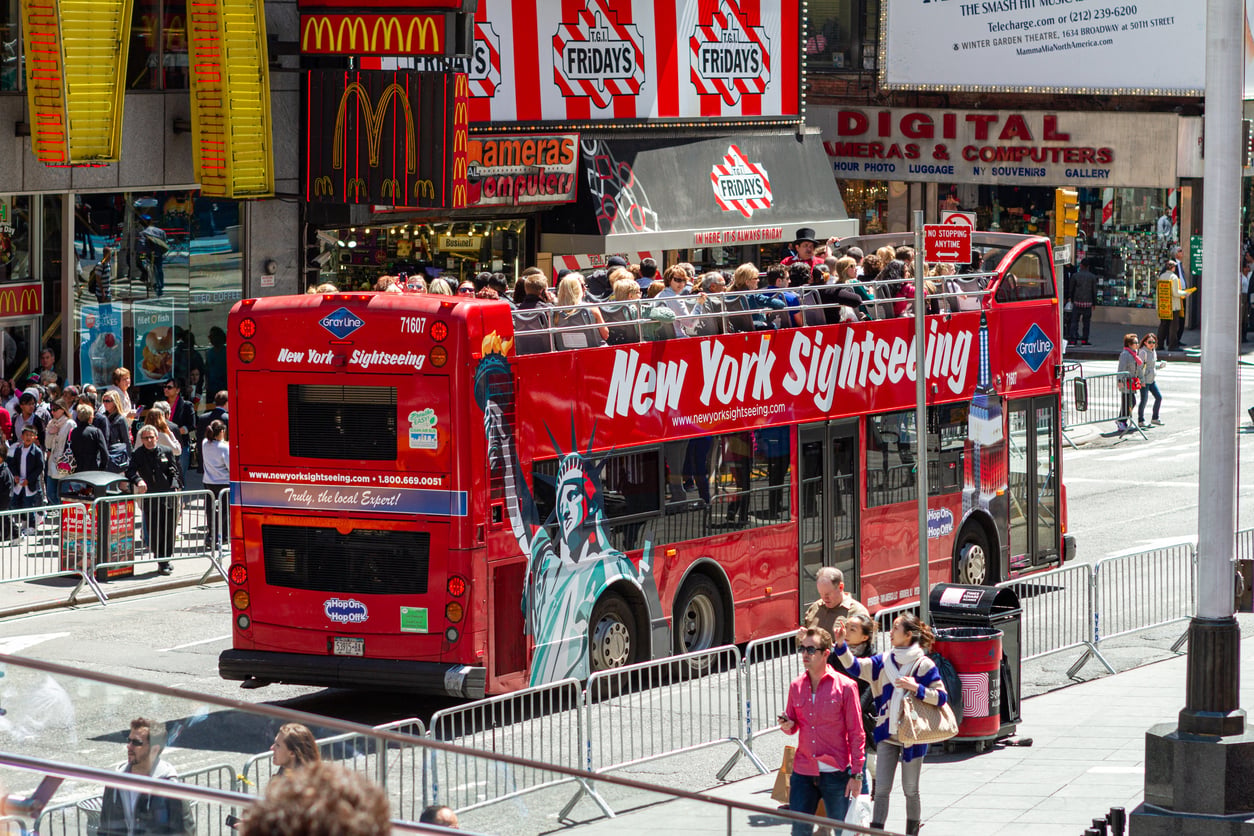 A red double-decker New York Sightseeing bus with tourists on the upper deck drives past McDonald's and TGI Friday's in a busy city area, surrounded by some of the city's most popular tourist attractions.