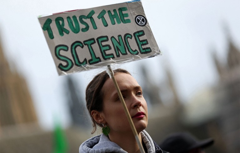 A protester holding a cardboard sign reading "TRUST THE SCIENCE", with an Extinction Rebellion logo.