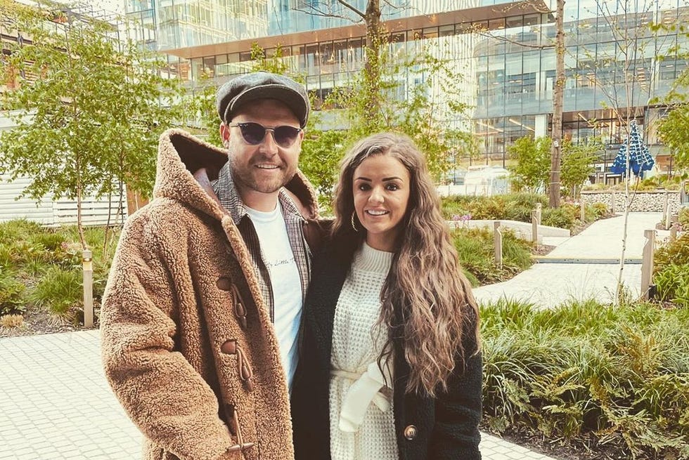 danny miller with fiancee steph jones posing in front of trees and glass building danny wears check trousers, a long furry coat, flat cap and sunglasses, while steph wears a white knitted dress and long black furry coat