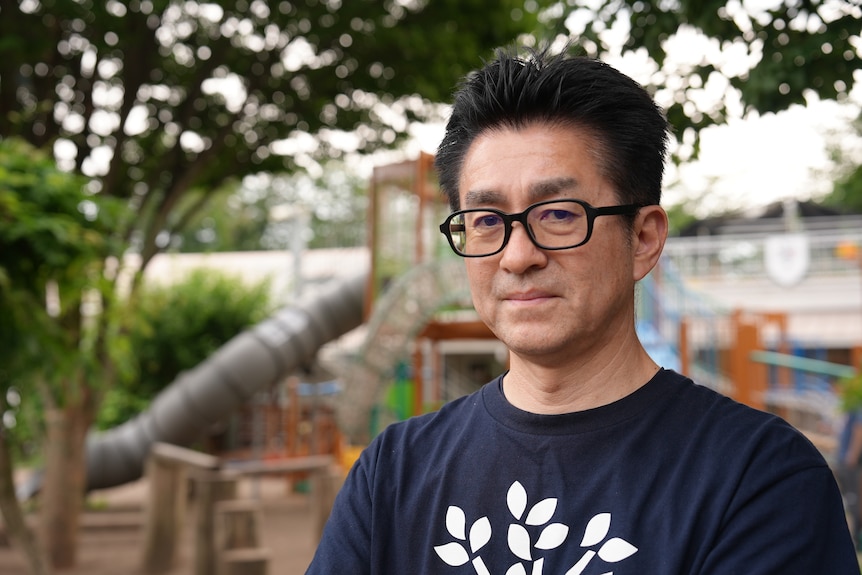 A man with black hair and glasses wearing a blue t-shirt with a children's playground blurred in background