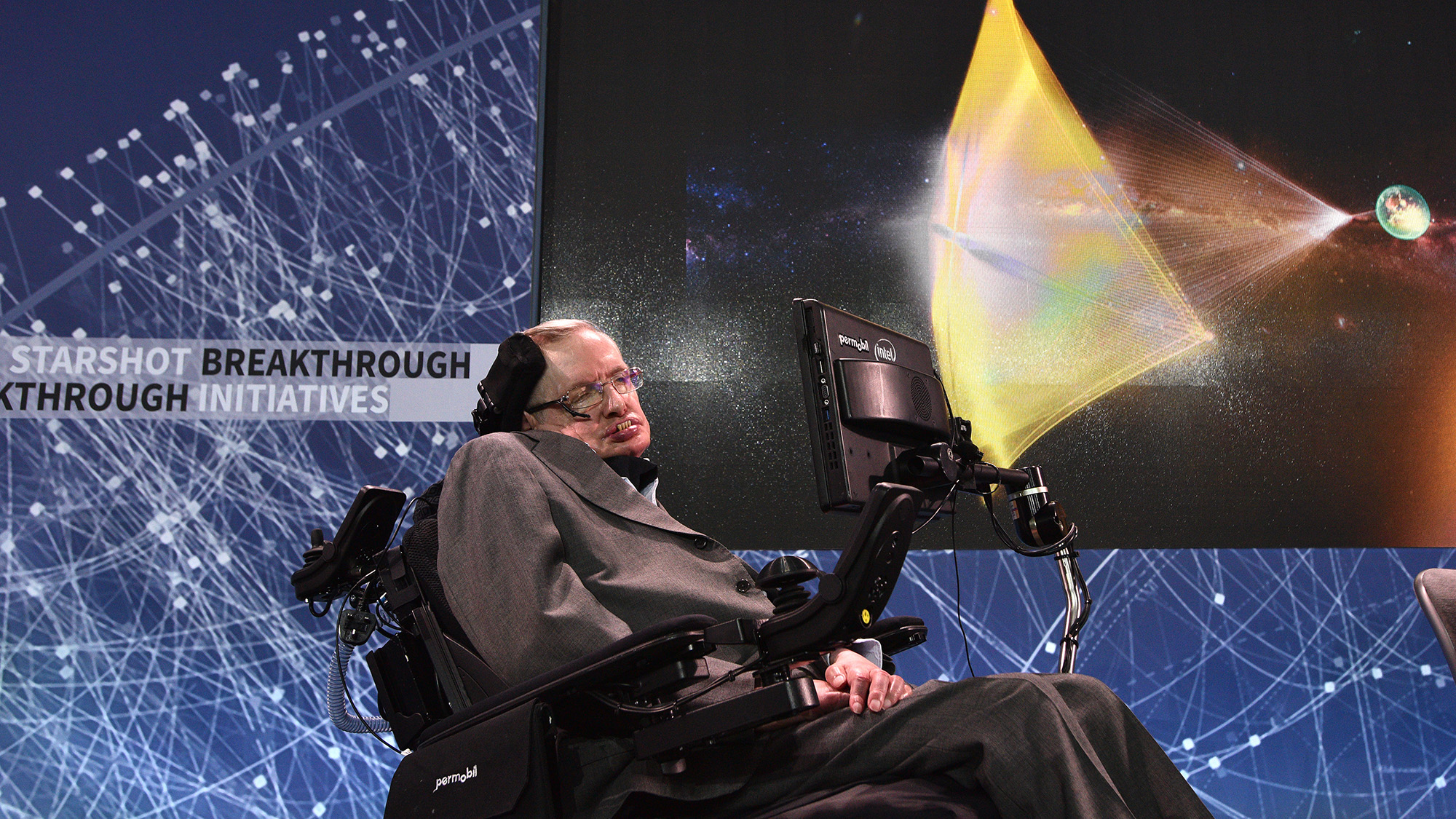 A man in an electronic wheelchair stands in front of a projector screen with various space images on it