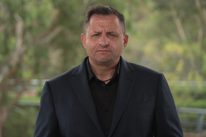 A man with short black hair wears a black jacket over a black collared shirt and stands in front of green trees.