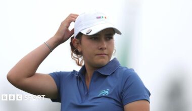 Charlotte Naughton, wearing a blue polo neck shirt, touches the back of her white baseball cap while playing for Great Britain and Ireland in the Curtis Cup
