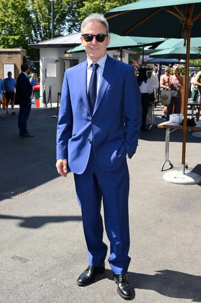 Jason Isaacs in a blue suit and tie, standing outside with umbrellas and people in the background.
